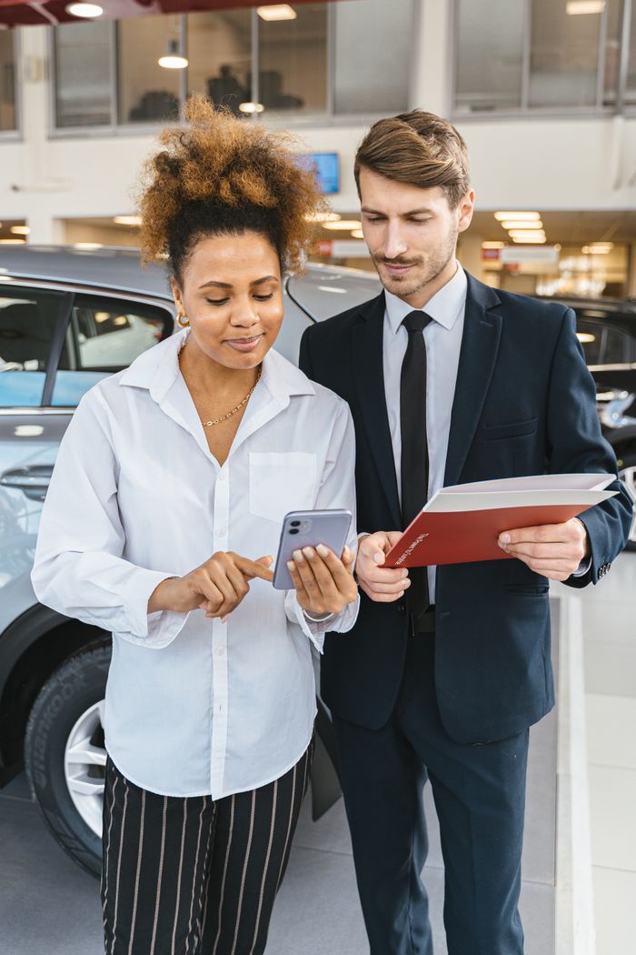 A man and woman reviewing car options on a mobile phone in a dealership showroom.